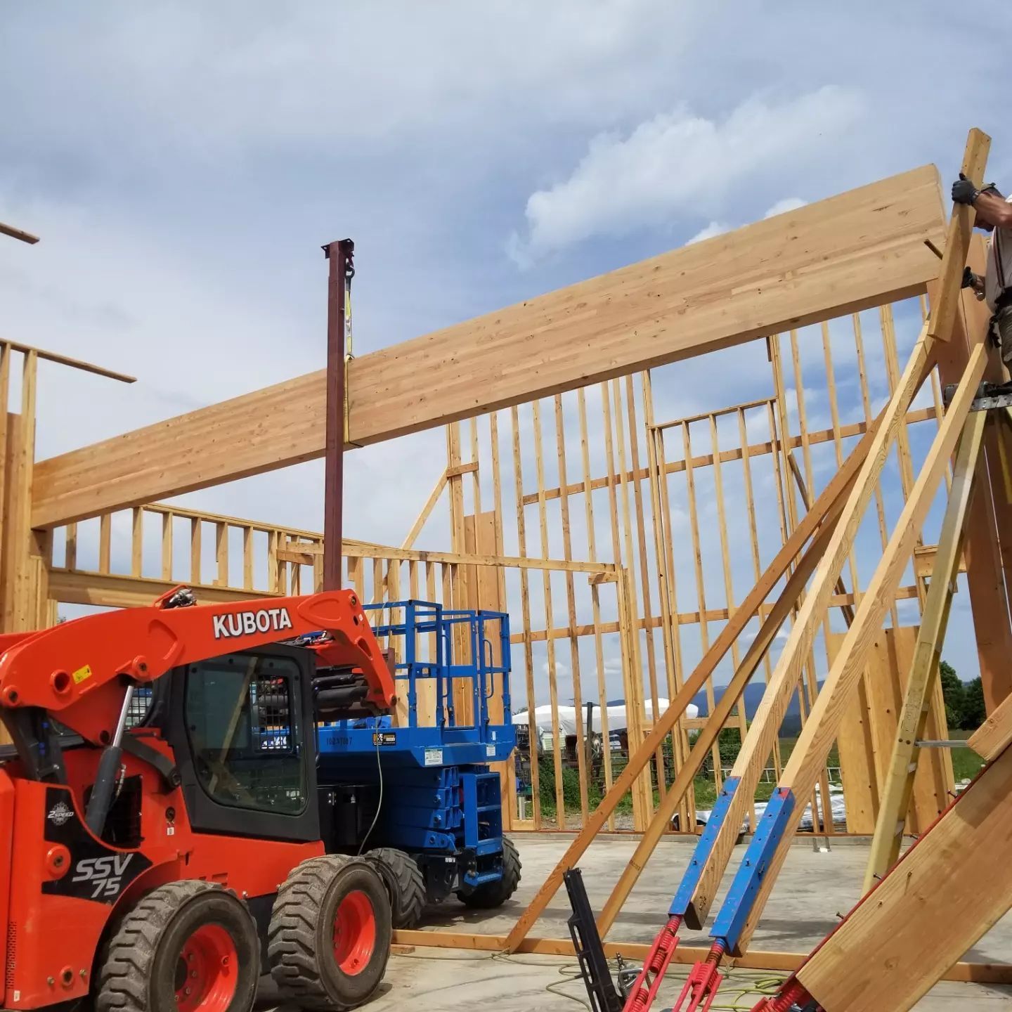Construction site with a Kubota skid steer, a blue lift, and a worker securing a wooden beam.