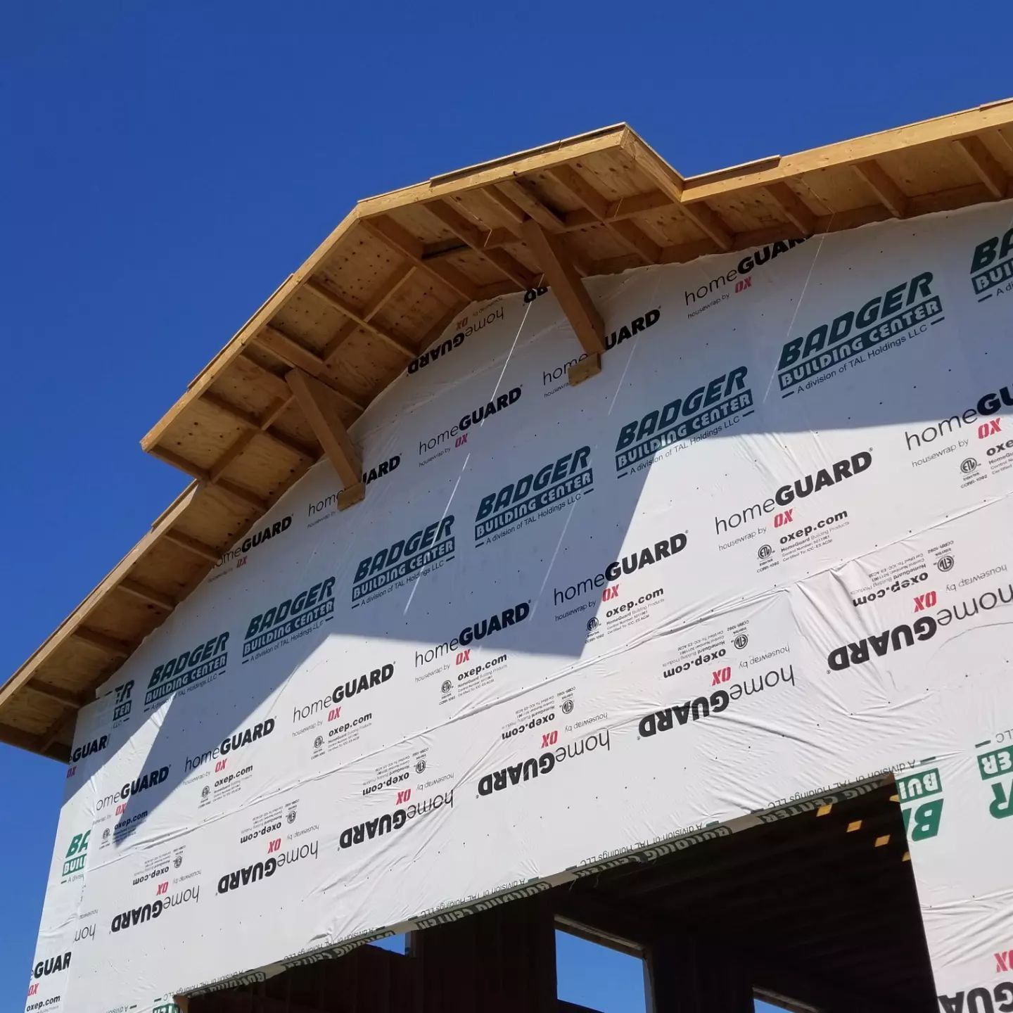 Roof of a house under construction with wood framing, white weather barrier, and blue sky.