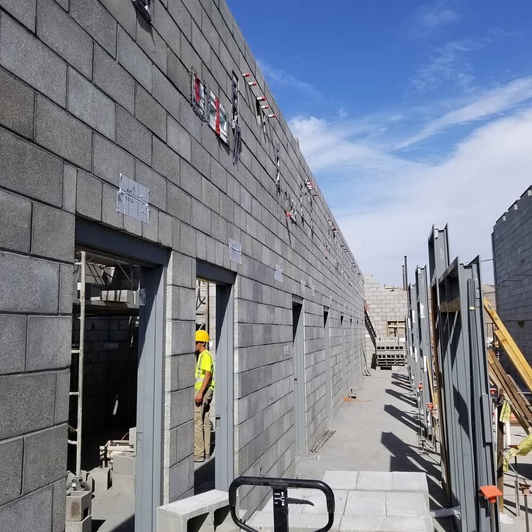 Exterior of a building under construction, grey block walls, steel doorways, a worker in a yellow hardhat is visible.