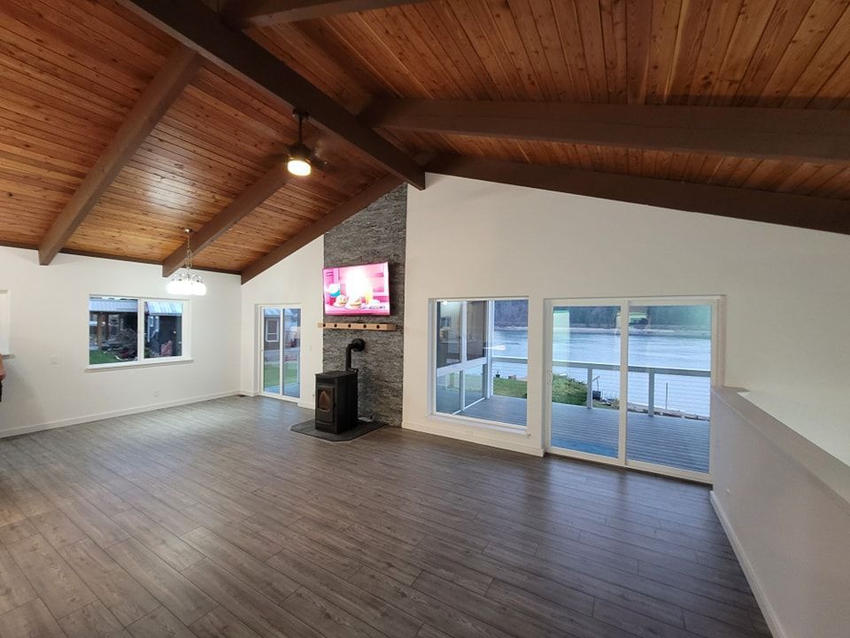 Spacious living room with vaulted wood ceiling, fireplace, large windows overlooking a body of water.