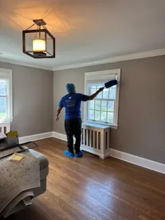 Person dusting window trim in a room with hardwood floors and light gray walls.