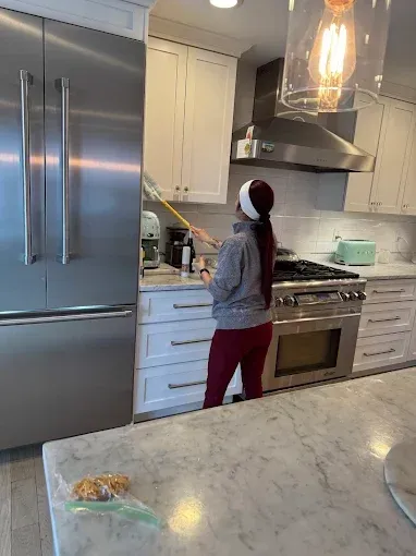 Woman dusting kitchen cabinets. Kitchen features stainless steel appliances, white cabinets, and marble countertops.