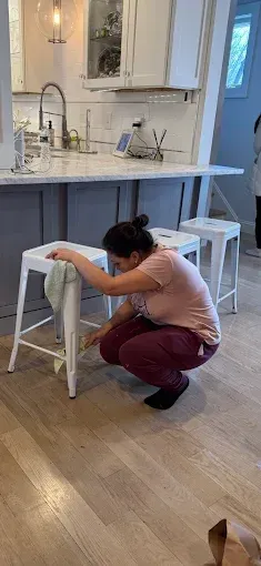 Woman cleaning a white bar stool in a kitchen with a gray island and wood floors.