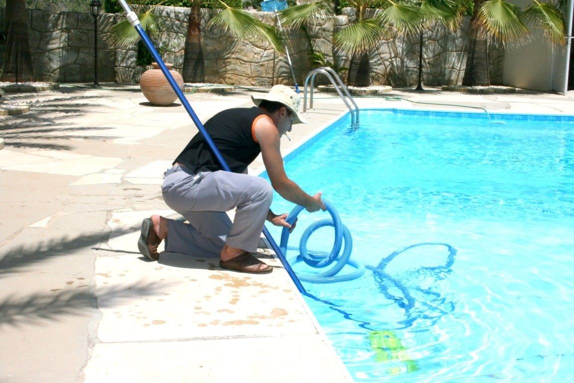 Person in a cowboy hat using a vacuum hose to clean a blue swimming pool in an outdoor setting.