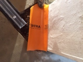 Orange Schluter Ditra membrane being installed near a drain in a light-colored setting.