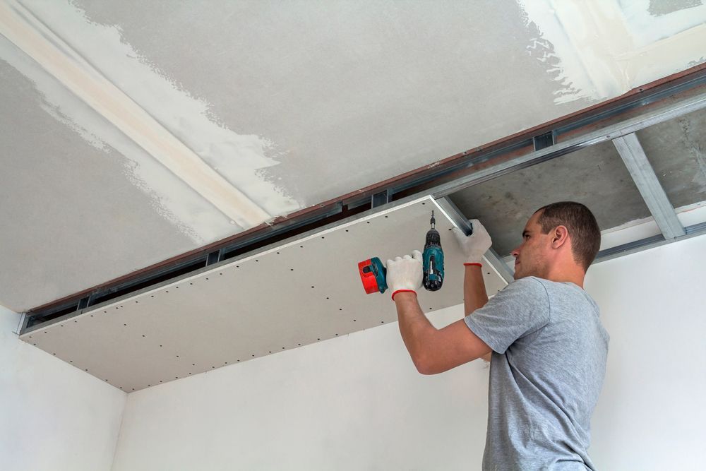 Man in gloves installing drywall on a ceiling with a drill.