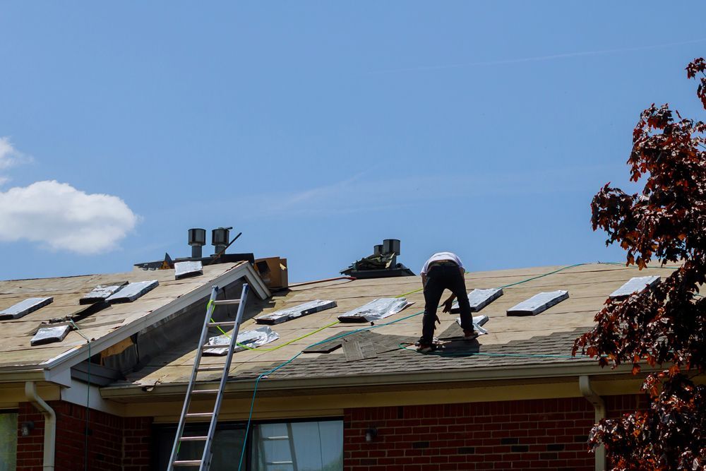 Roofer working on a partially shingled roof with a ladder, on a sunny day.