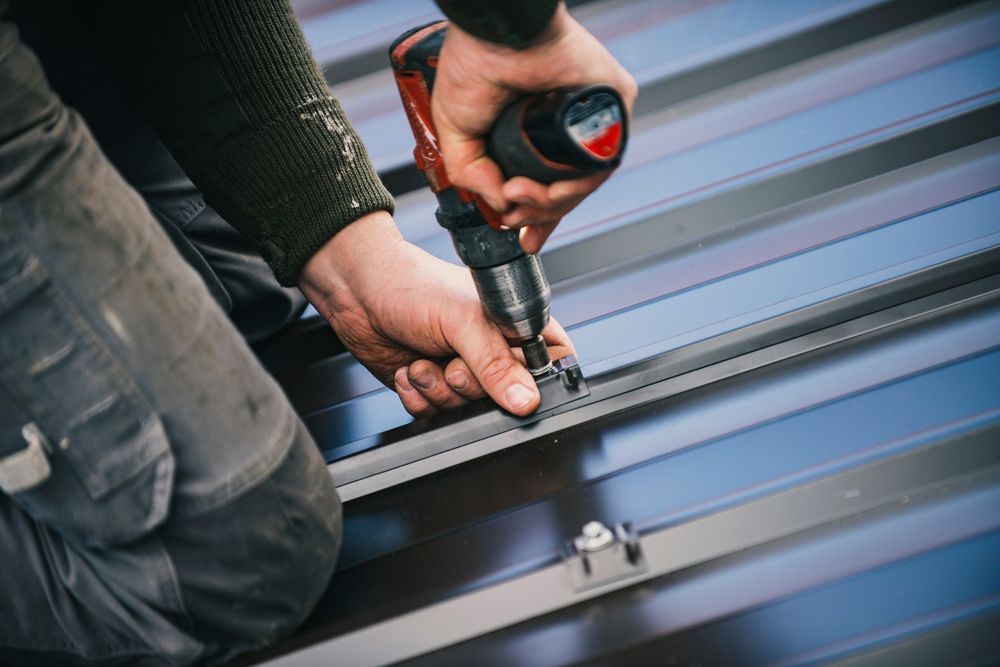 Person using a power drill to install metal roofing. Close-up view shows hands, drill, and roof panels.