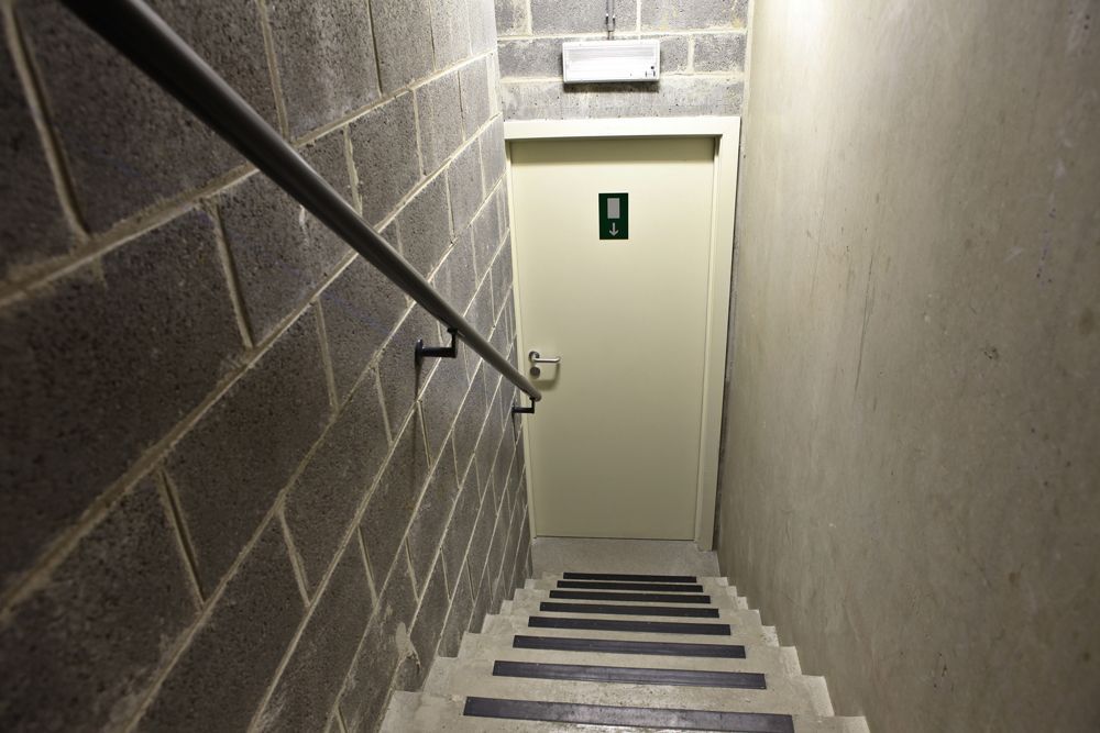 Stairwell descending towards a door with a restroom sign, gray block walls, metal railing, and dim lighting.