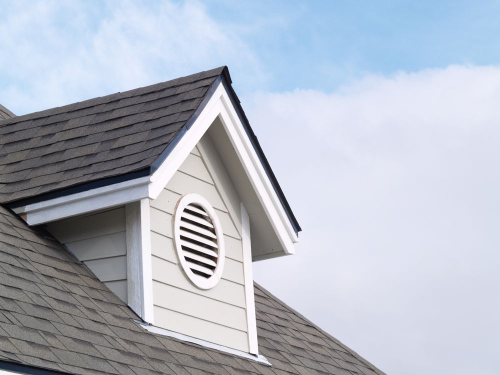 Dormer with oval vent on a roof against a partly cloudy sky.