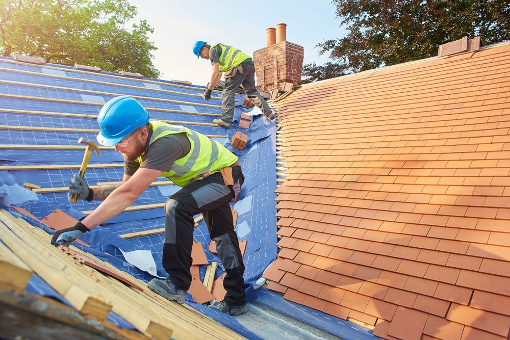 Two roofers in hard hats, hammering shingles on a roof during the day.
