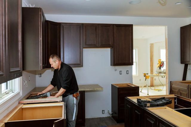 Man installing kitchen cabinets, dark brown against white walls. Partially built kitchen, unfinished.