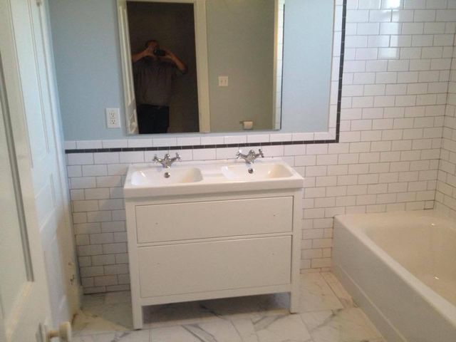White bathroom with double sink, tub, subway tile, and a large mirror reflecting a person.