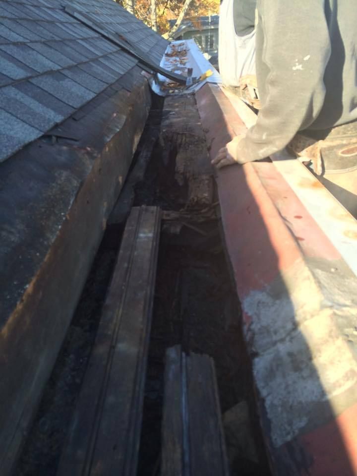 Person cleaning out debris from a gutter. Shingles, a red gutter, and wood visible.