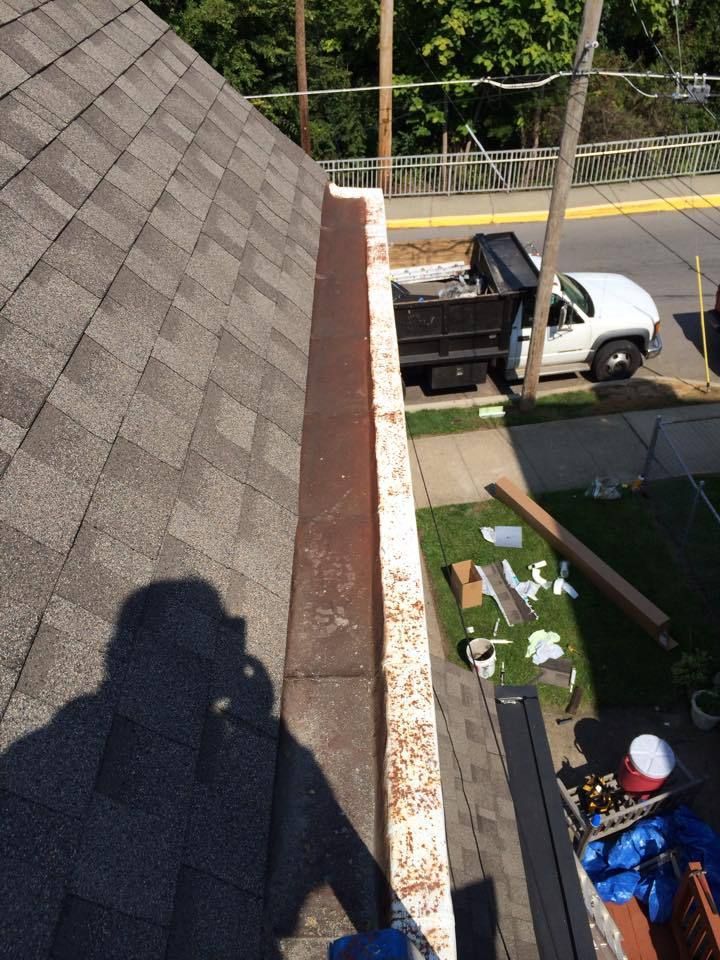Rooftop view showing a rusty gutter, asphalt shingles, and a truck on a street with tools, supplies, and a worker’s shadow.