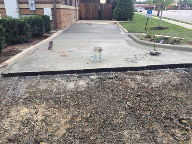 Concrete driveway under construction; exposed rebar, fresh pour, bucket, curb, building, outdoors.