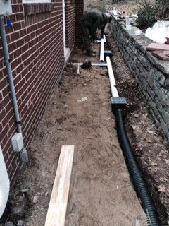 Man installing drainage pipes along a brick house, next to a stone wall.