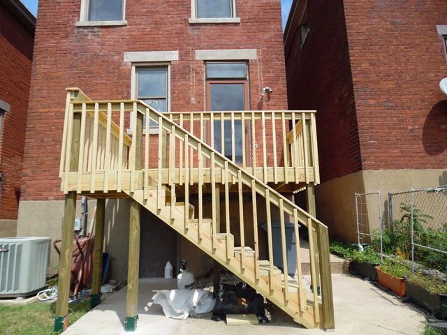 Wooden deck with stairs attached to a brick building. Green support beams and railing are visible.