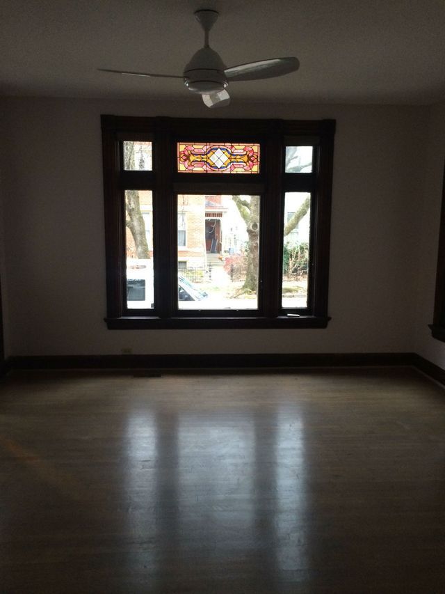Empty room with a large window, stained glass, and a ceiling fan. Wooden floors reflect light.
