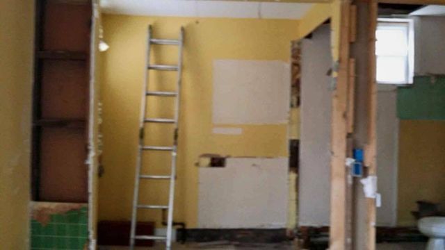 Bathroom undergoing renovation; yellow walls, ladder, exposed framing, and partial tile.