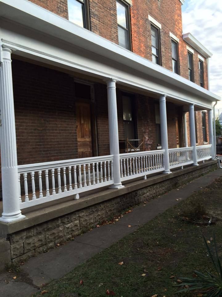 White-columned porch on brick building. White railing, gray stone base, and green grass in front.