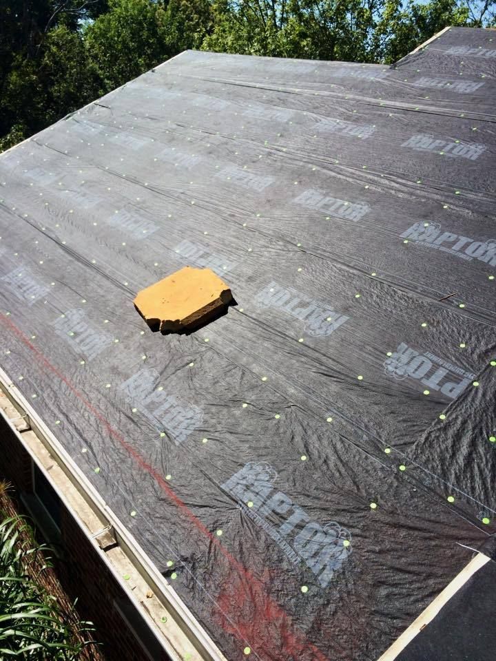 Black roofing underlayment on a roof, with a yellow, octagonal object in the center. Green trees in background.