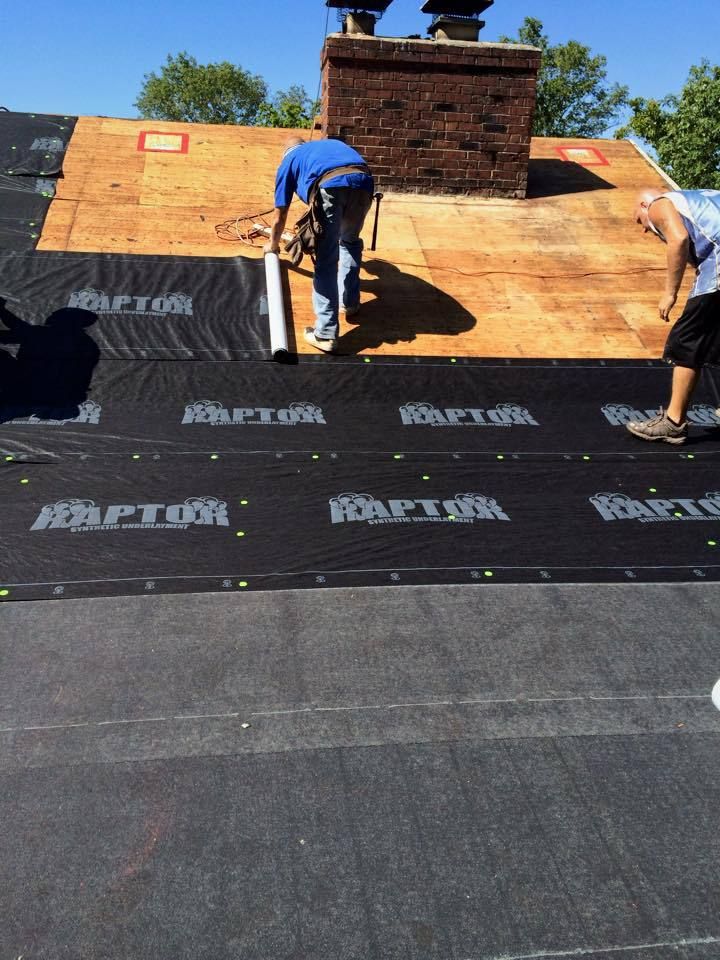 Two roofers installing black underlayment on a roof near a brick chimney on a sunny day.