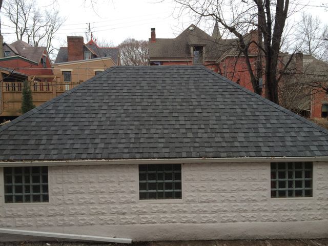 A white building with a dark grey shingle roof, three square windows, and red brick houses in the background.