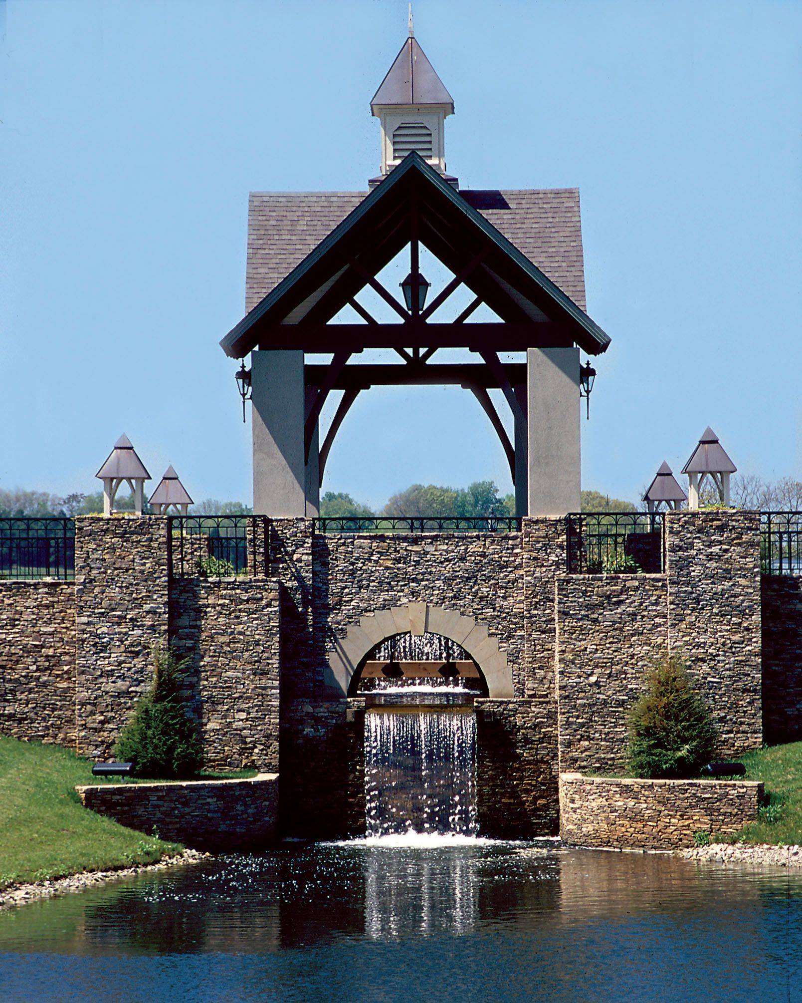 Stone and wood structure with a waterfall cascading into a pond, under a blue sky.
