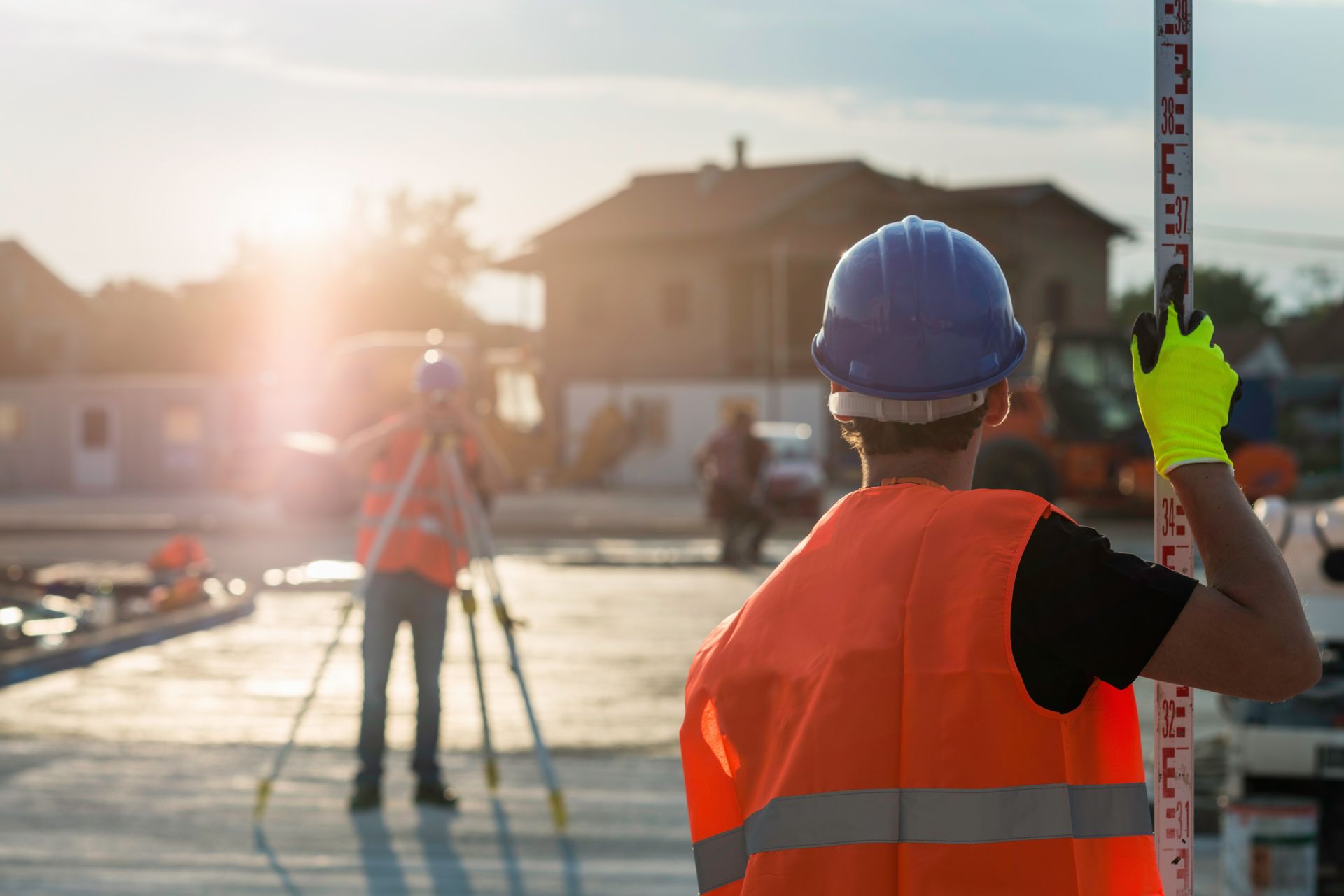 Worker holding a leveling rod as another surveys a residential construction site at sunset. Worker holding a leveling rod as another surveys a residential construction site at sunset.