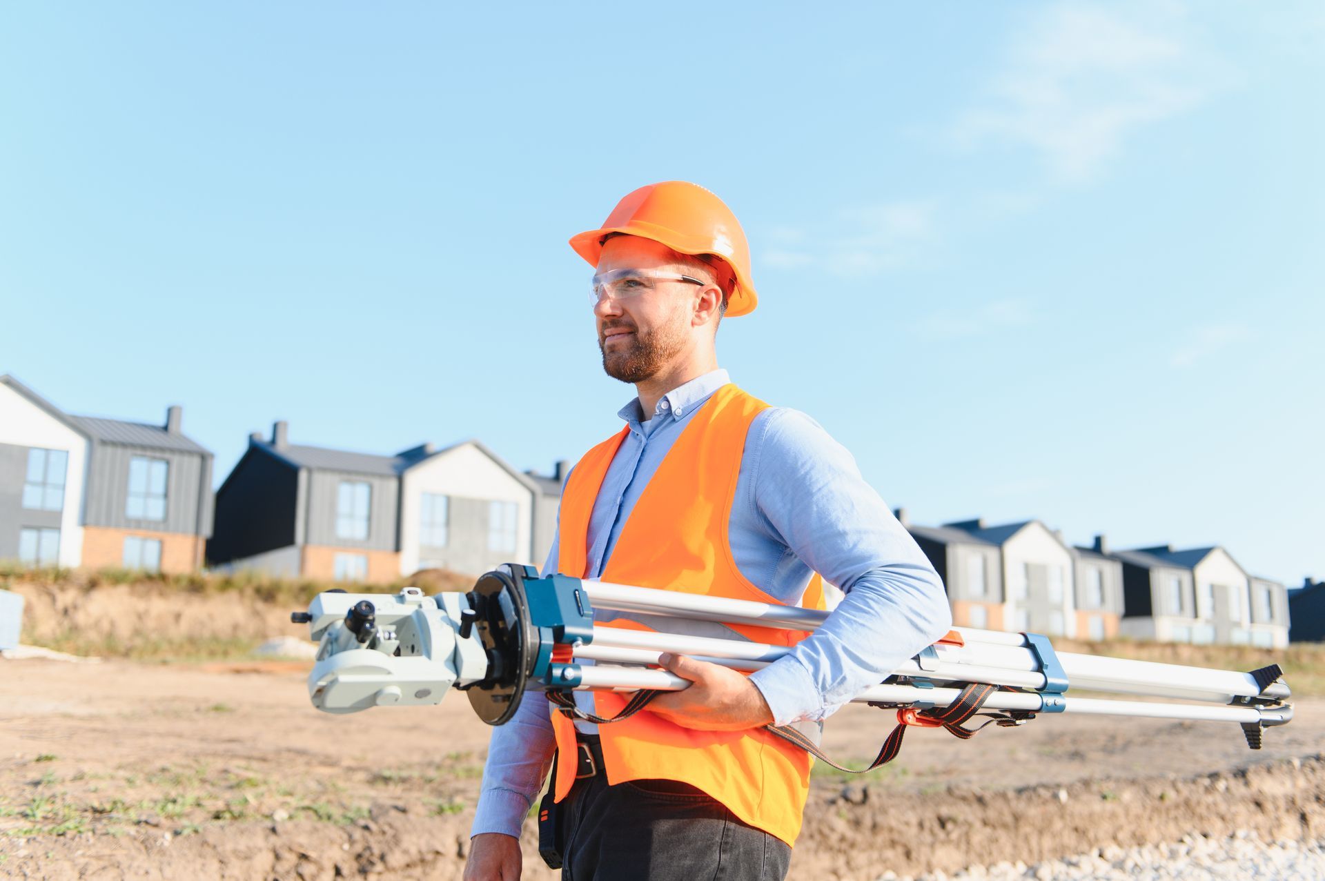 Worker carrying surveying equipment at a housing site with new homes in the background. Worker carrying surveying equipment at a housing site with new homes in the background.
