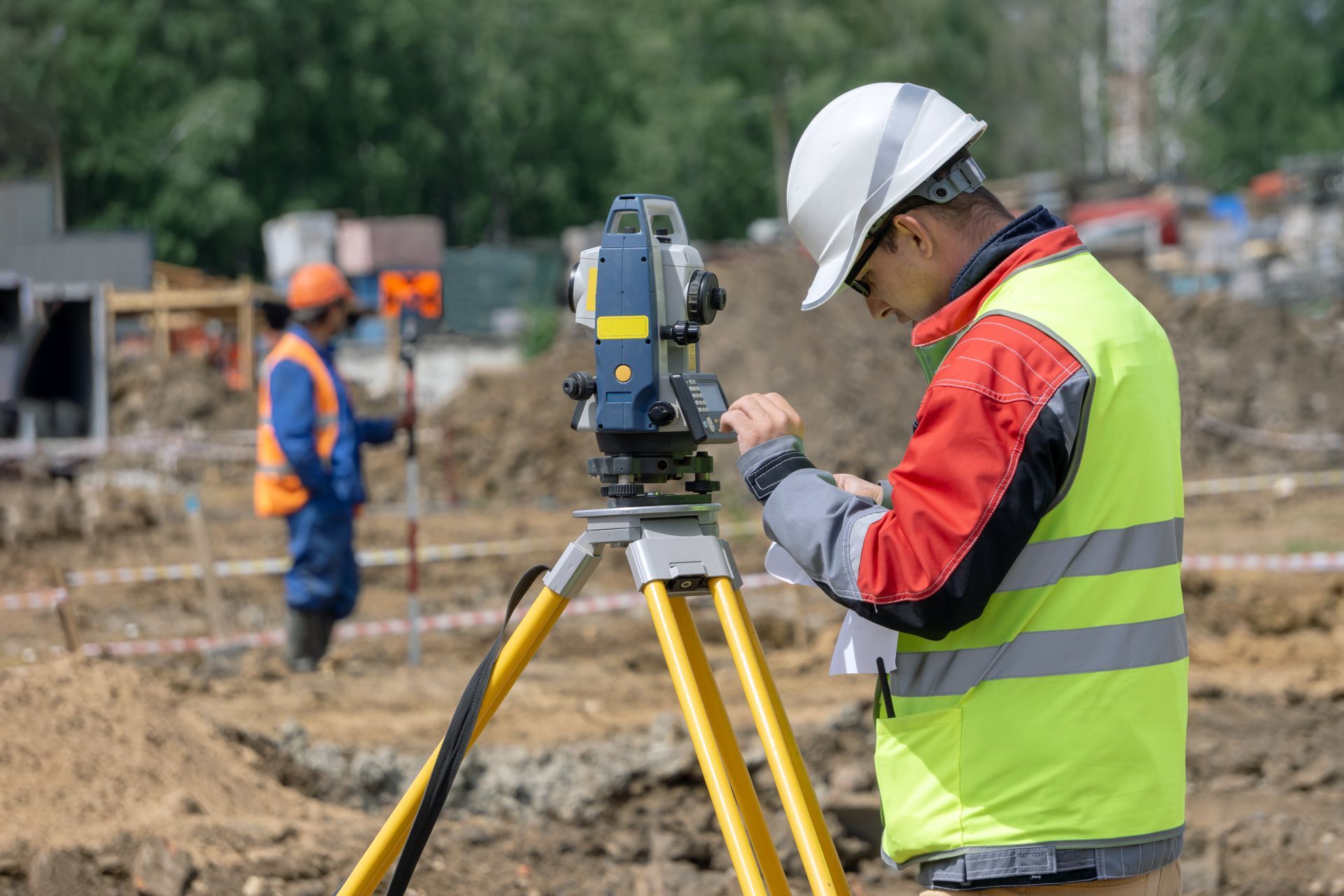 Surveyor records measurements while using a tripod-mounted total station on an active jobsite. Surveyor records measurements while using a tripod-mounted total station on an active jobsite.