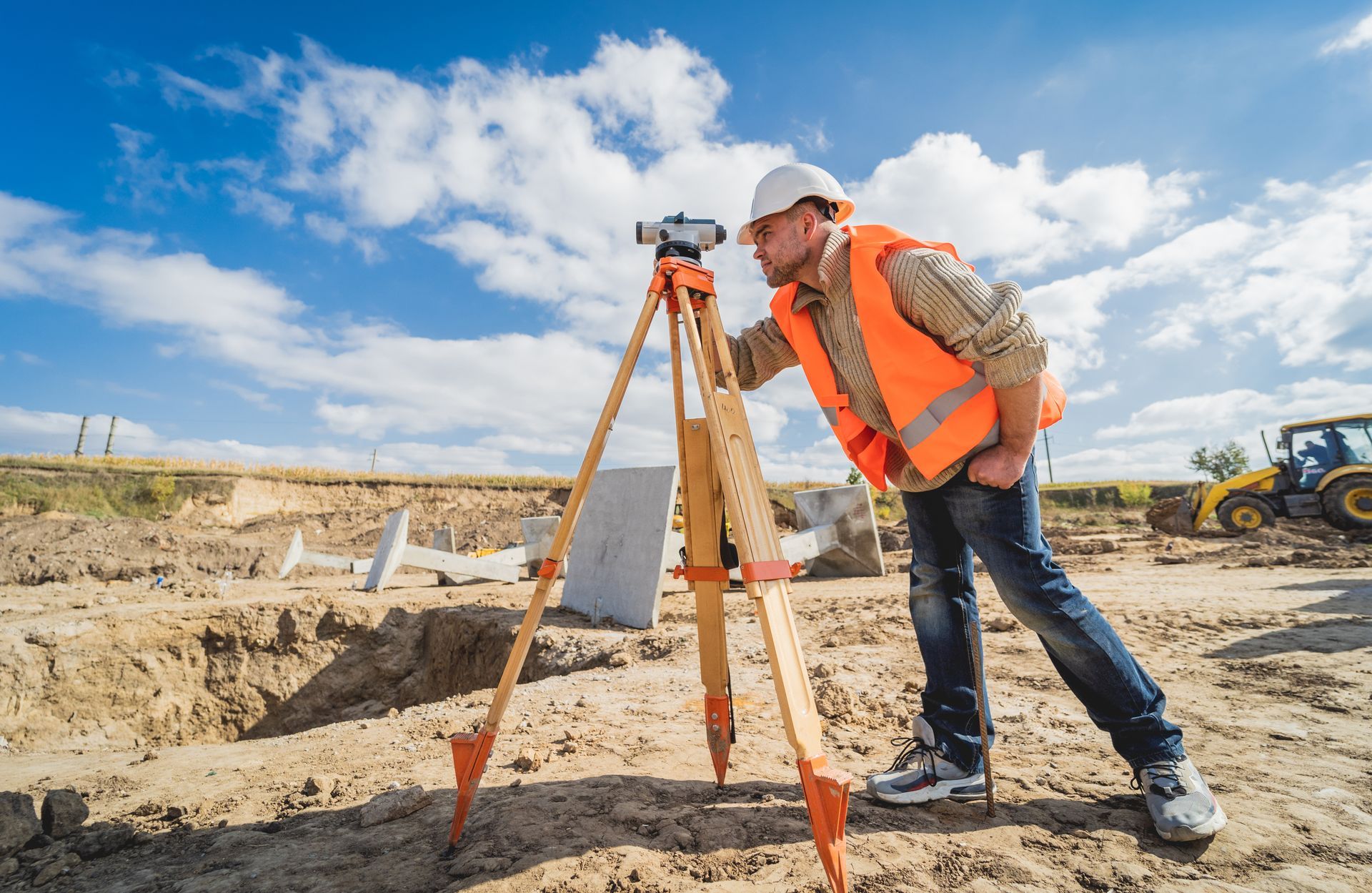Surveyor using tripod-mounted level at a construction site under a bright sky.