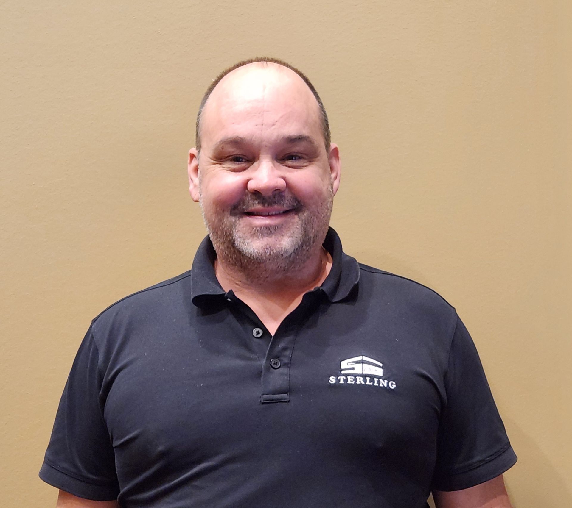 A middle-aged man with a bald head and goatee wearing a black polo shirt smiles against a tan wall.