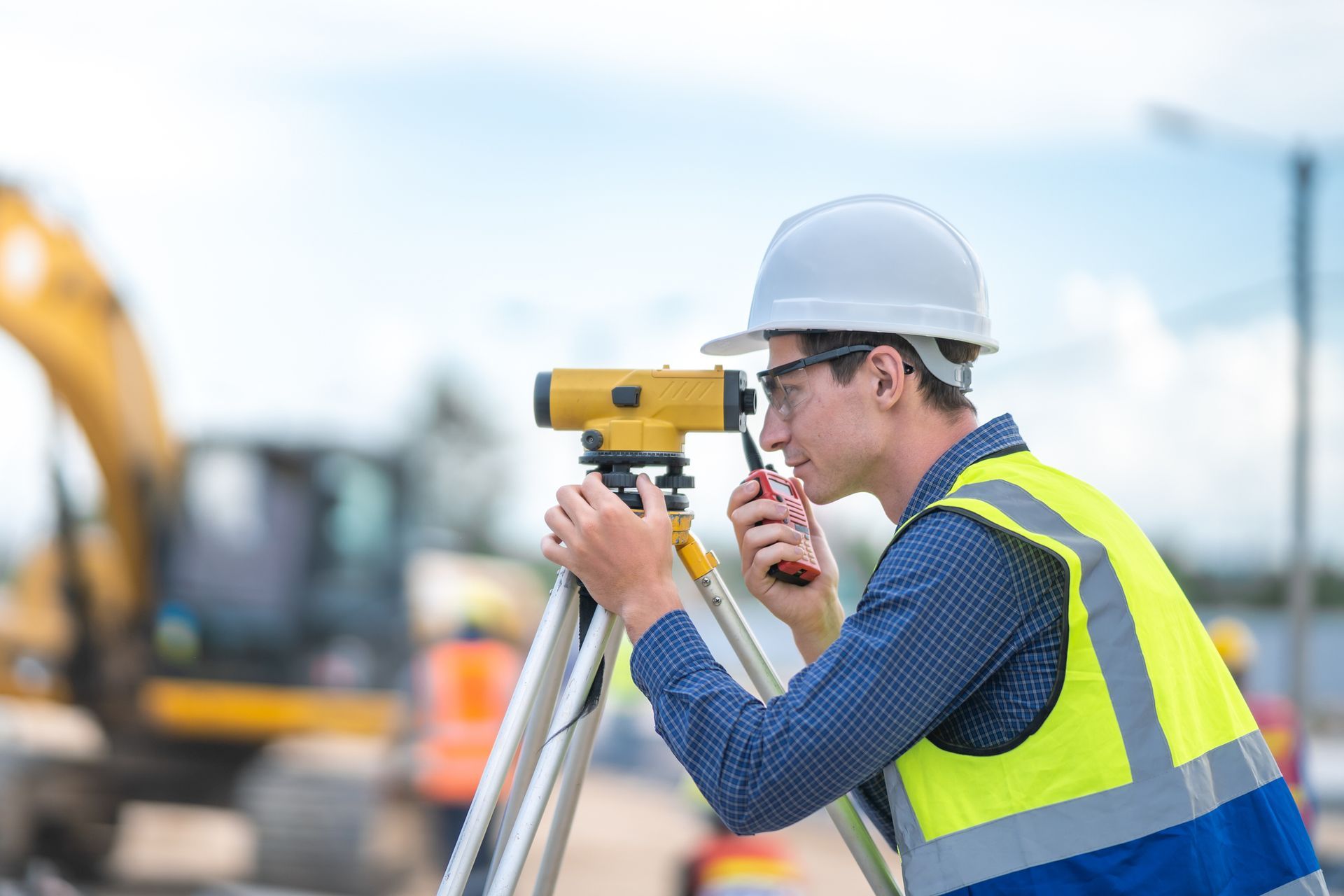Construction worker in hard hat and safety vest using a surveying level outdoors, with construction equipment in the blurred background.