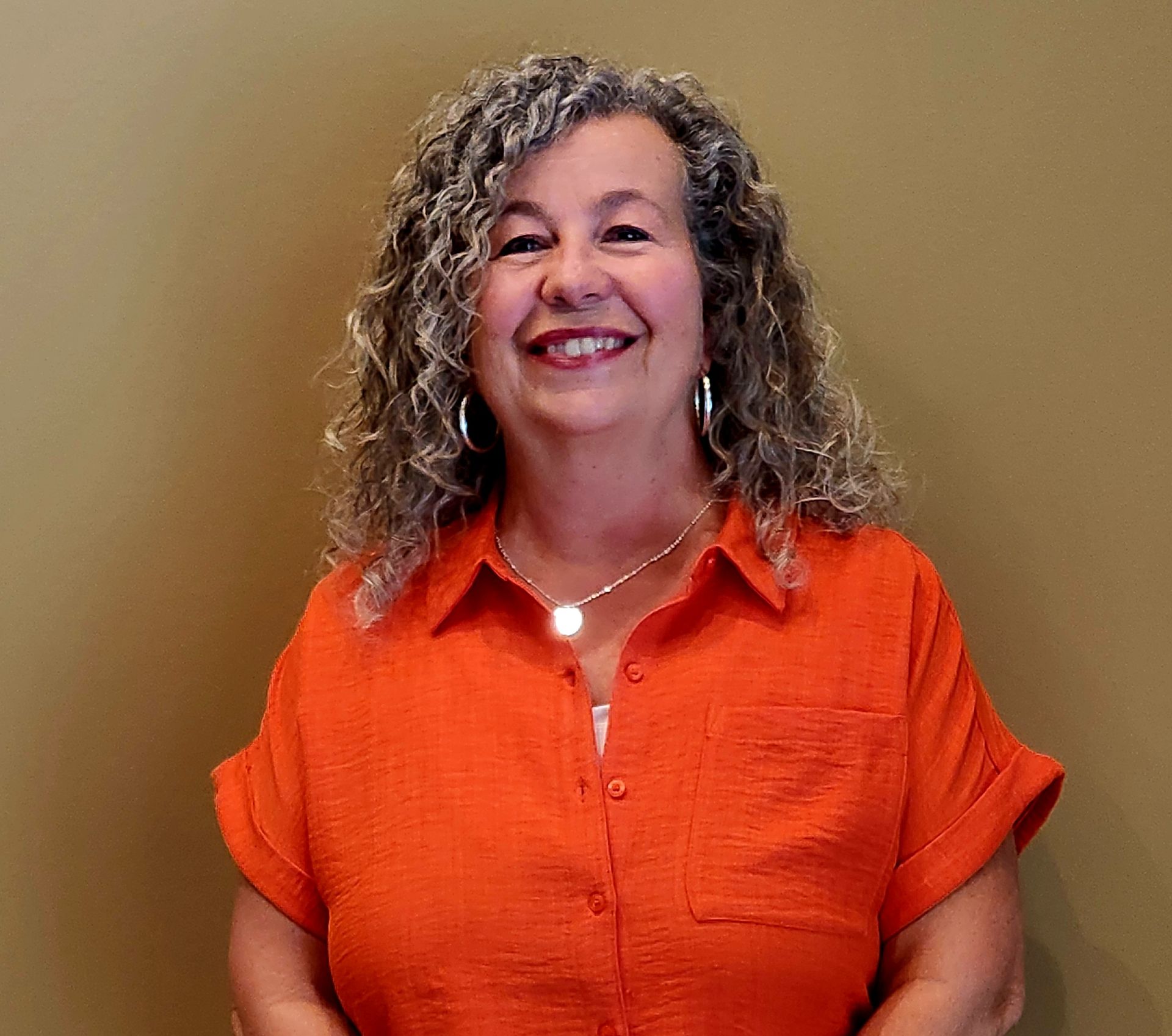 Woman with curly gray hair smiles, wearing an orange button-down shirt and necklace against a tan wall.