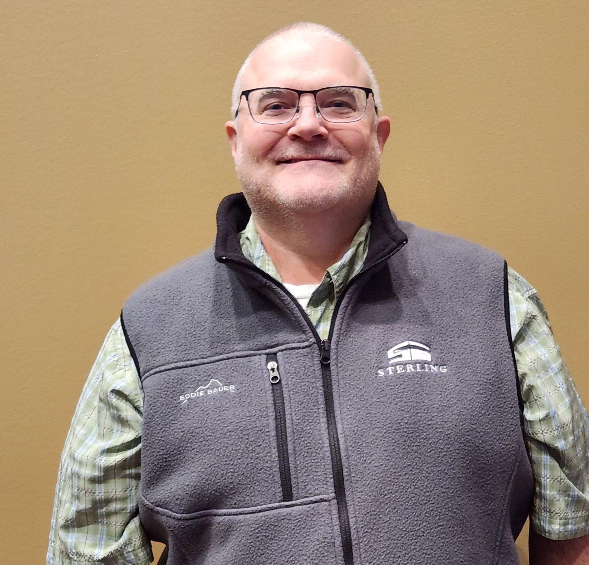 A smiling, older man with glasses wearing a gray vest in front of a tan wall.