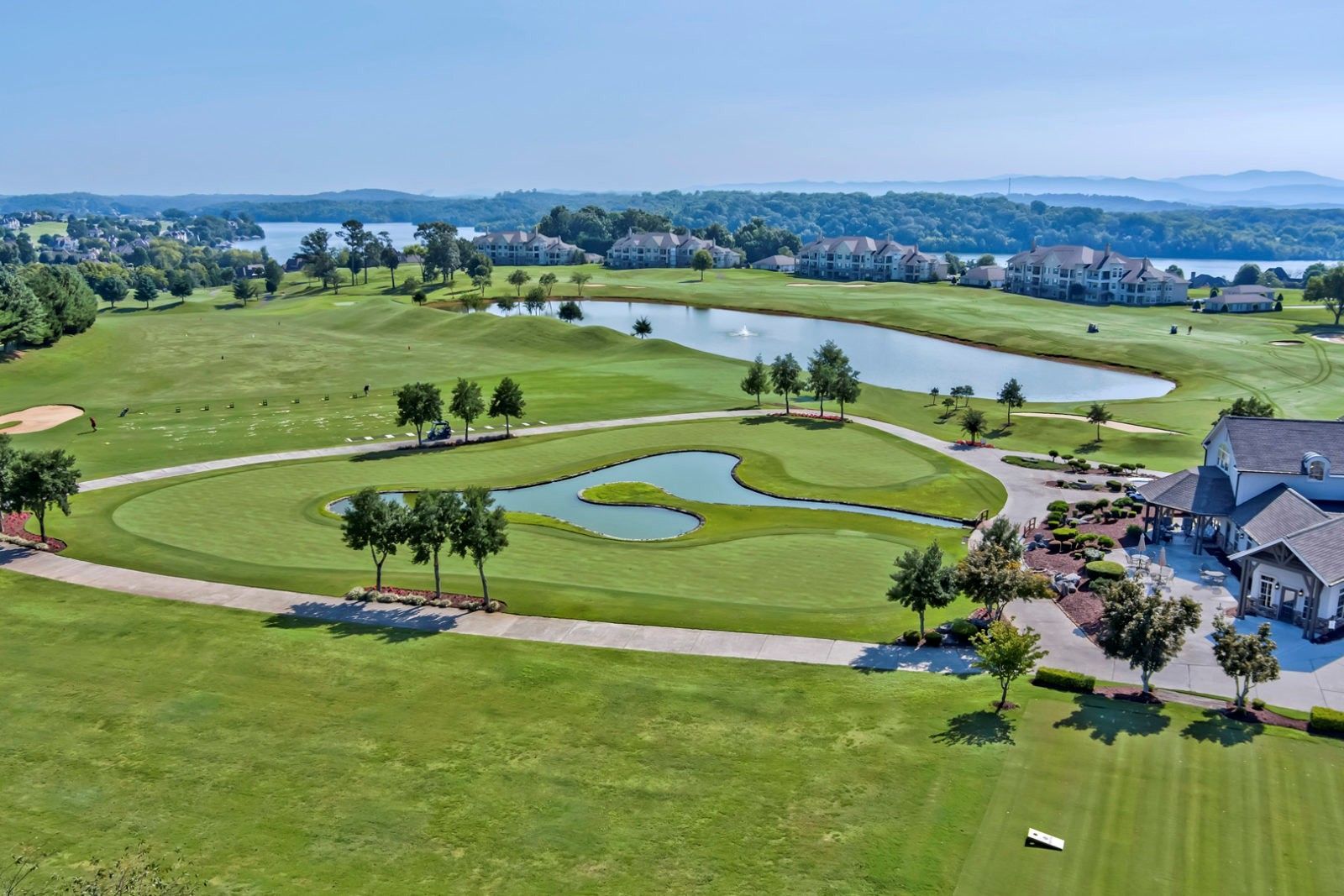 Aerial view of a golf course with a large green, water features, and a clubhouse in the foreground. A lake and houses are in the background.