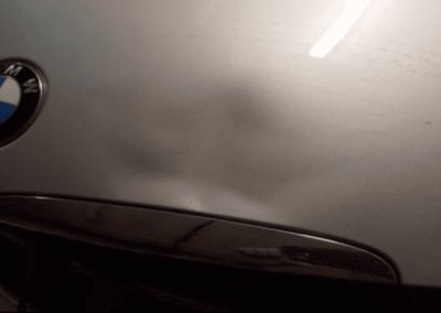 Silver BMW hood with emblem and dark, possibly damaged, area.