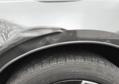 Black car's dented rear fender above the tire, with a blurry background of trees.