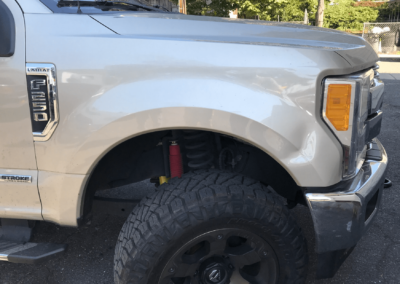 Tan Ford F-350 truck front quarter panel. Black tire, silver fender, red shock absorber, yellow spring.