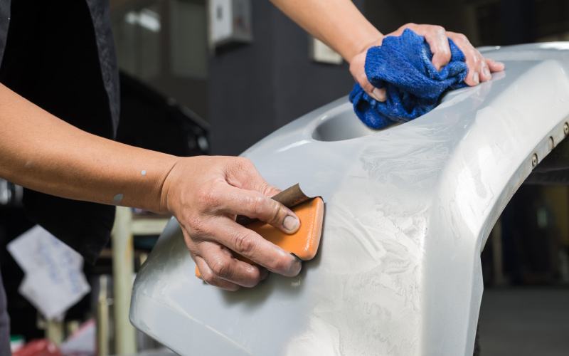 Person sanding a gray car bumper with an orange sanding block; blue cloth in background.