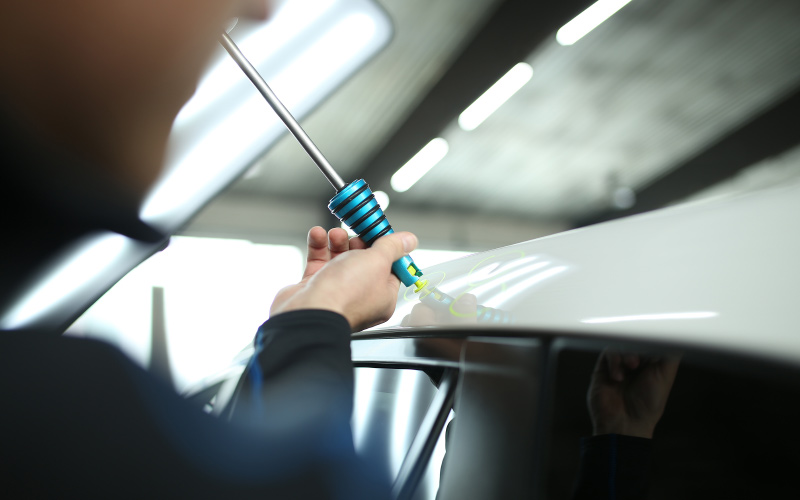 A person using a specialized tool to remove a dent from a white car's body in a repair shop.