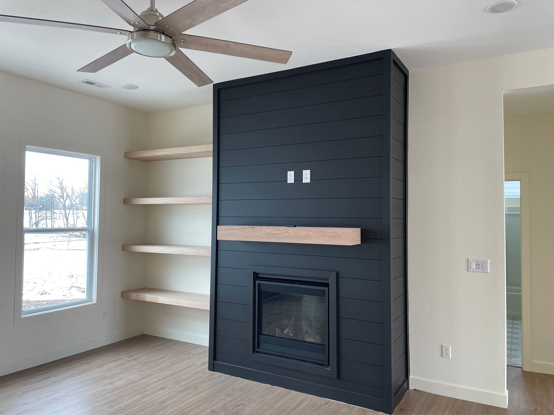 A modern living room with a black fireplace, light-colored shelves, and a ceiling fan.
