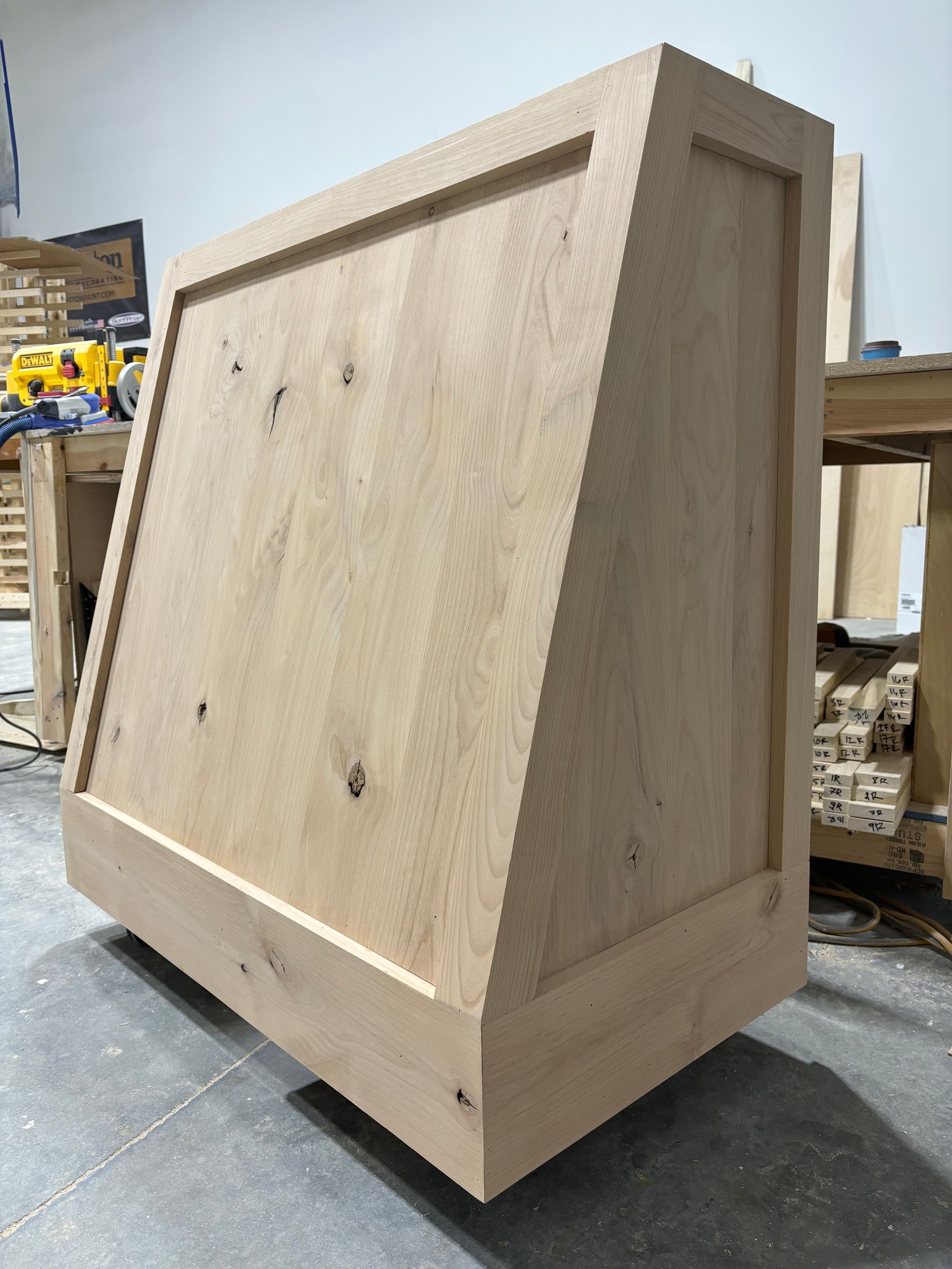 Wooden range hood in a workshop, unfinished and angled, with a rectangular trim around the face.