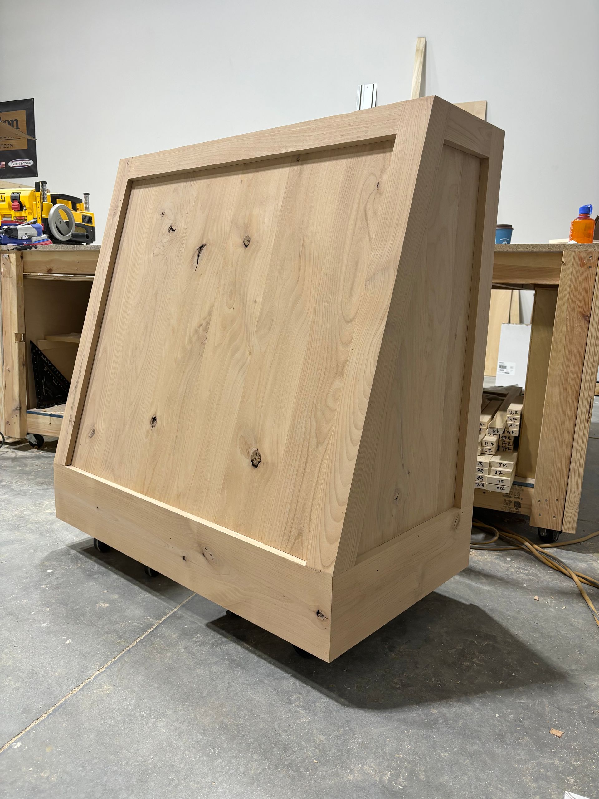 Angled wooden cabinet, unfinished, on a concrete floor in a workshop.
