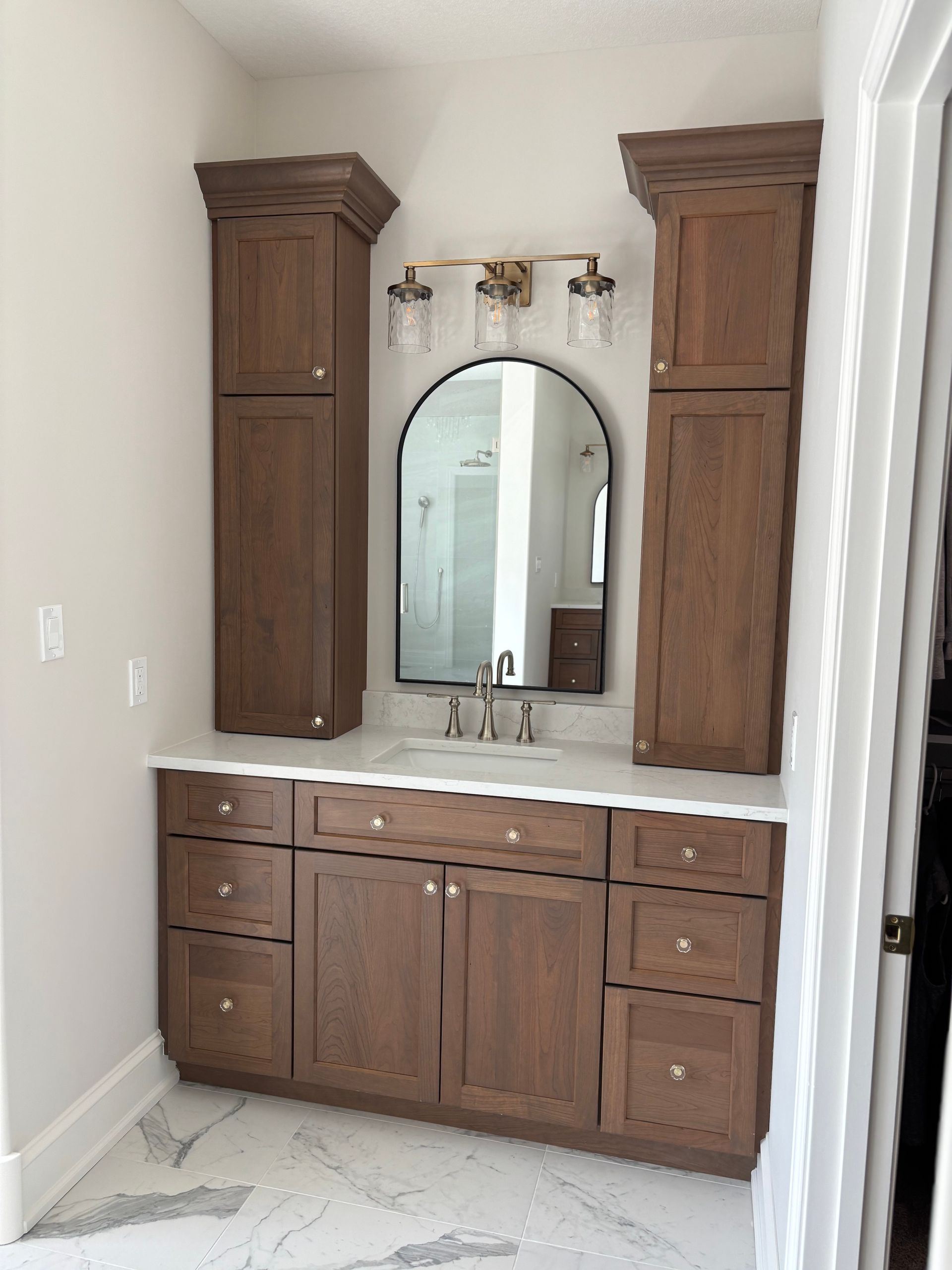 Bathroom vanity with brown cabinets, white countertop, and arched mirror.