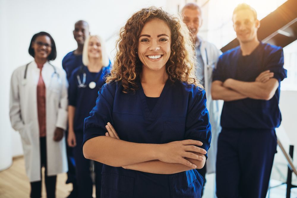 A diverse group of medical professionals in scrubs and white coats, smiling with arms crossed in a bright setting.