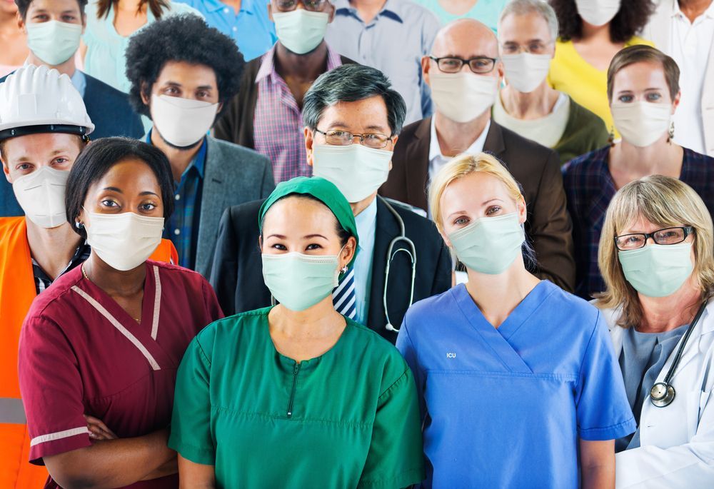 A diverse group of people wearing face masks, including medical staff, construction workers, and office workers, indoors.