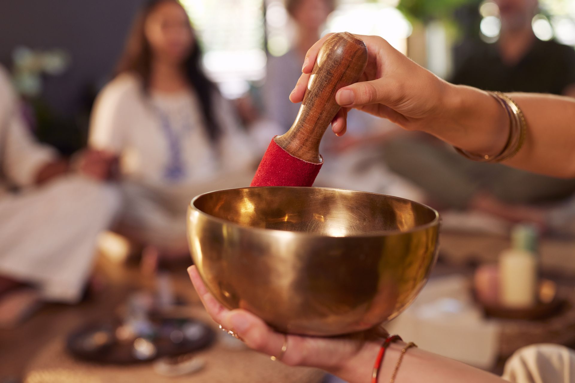 A person holds a golden singing bowl and mallet, with blurred participants in the background of a wellness setting.